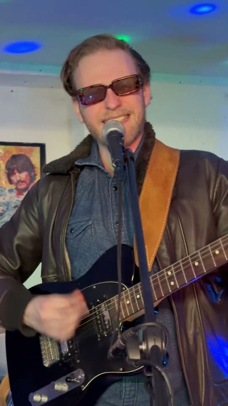 Sänger mit Telecaster am Mikrofon, dahinter ein Beatles-Bild an der Wand